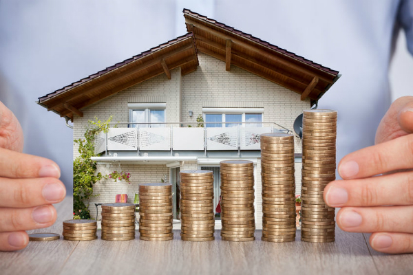 Real estate investment: Two hands (a man) with a house model on a table surrounded by stacks of gold-colored coins.